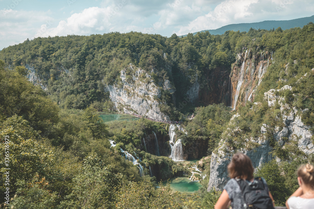 Group of tourists walking in Plitvice Lakes National Park on wood bridge in forest. Young tall blond girl carries her backpack on mountain path that leads to beautiful touristic Croatian places.