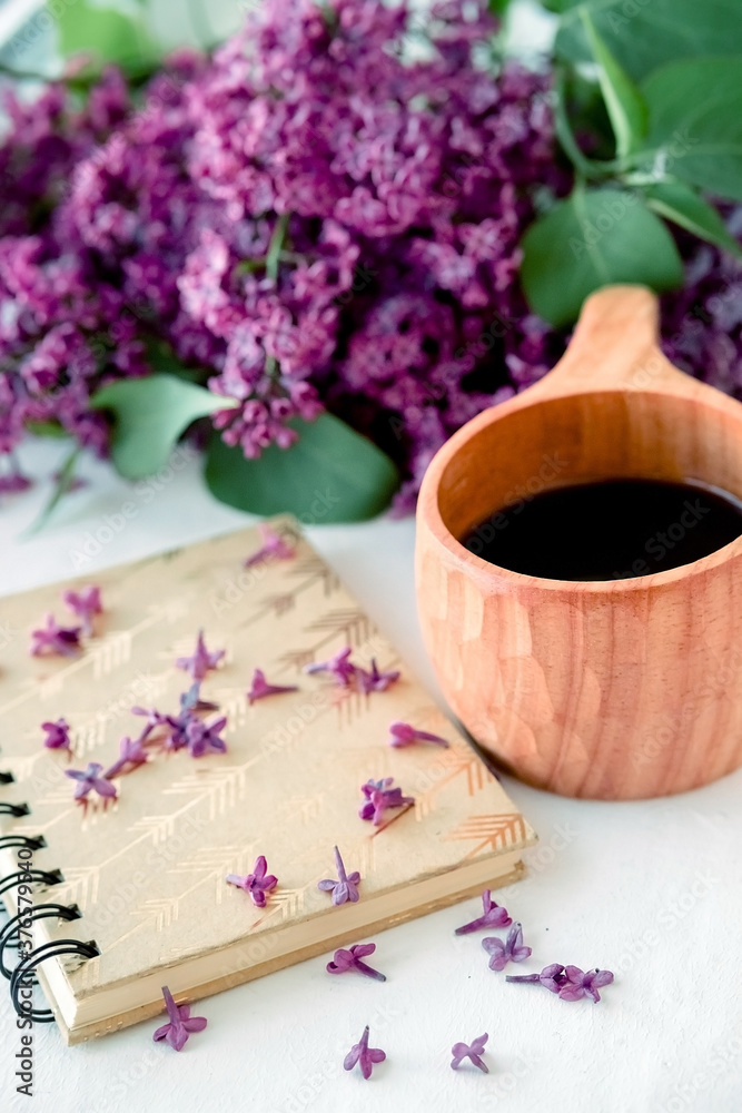 Wooden finnish cup with coffee on the table. Nearby lies a closed notebook for notes and a bouquet of lilacs. Good morning and good mood, planning a day at breakfast, concept.