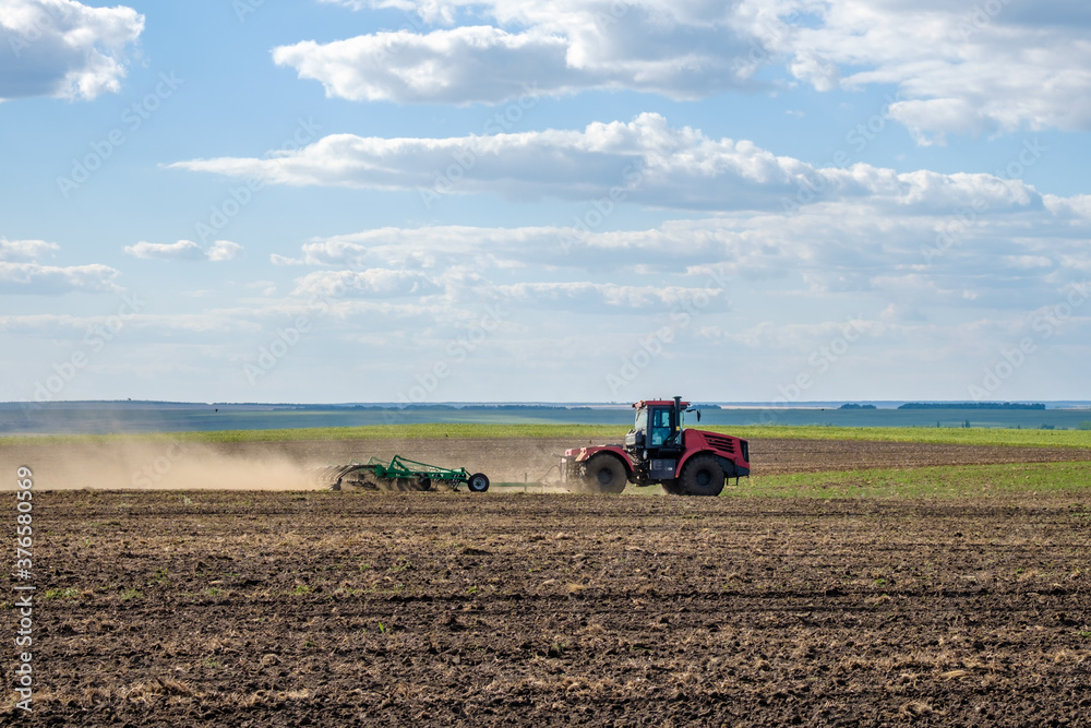Obraz premium A red farm tractor in a cloud of dust cultivates the soil in the field with a cultivator after harvest. Summer sunny day. Fertile land. Modern agricultural machinery. Copy space. High quality photo
