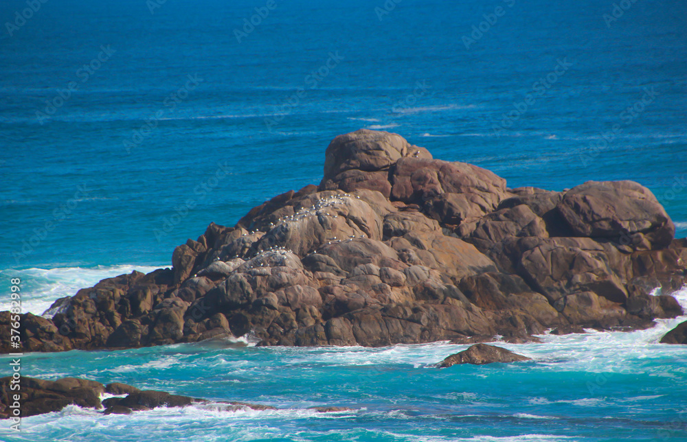 Scenic ancient Sugar Loaf Rock South Western Australia in the blue Indian Ocean is a popular fishing and hiking destination with its treeless green dunes and  splashing waves on old eroded rocks.