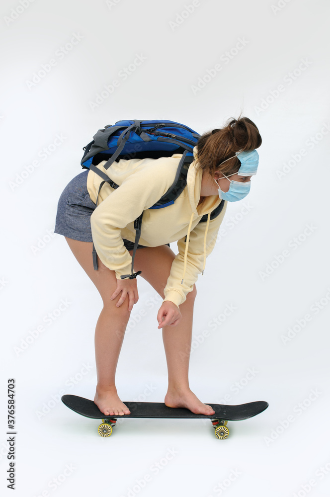 Naklejka premium Vertical photo of a barefoot teen girl with the masks on the face and a backpack rolling on the skate board over the white background