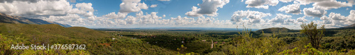 Panorama of forested mountains landscape on a sunny day with clouds shaping shadows over the houses of a near town