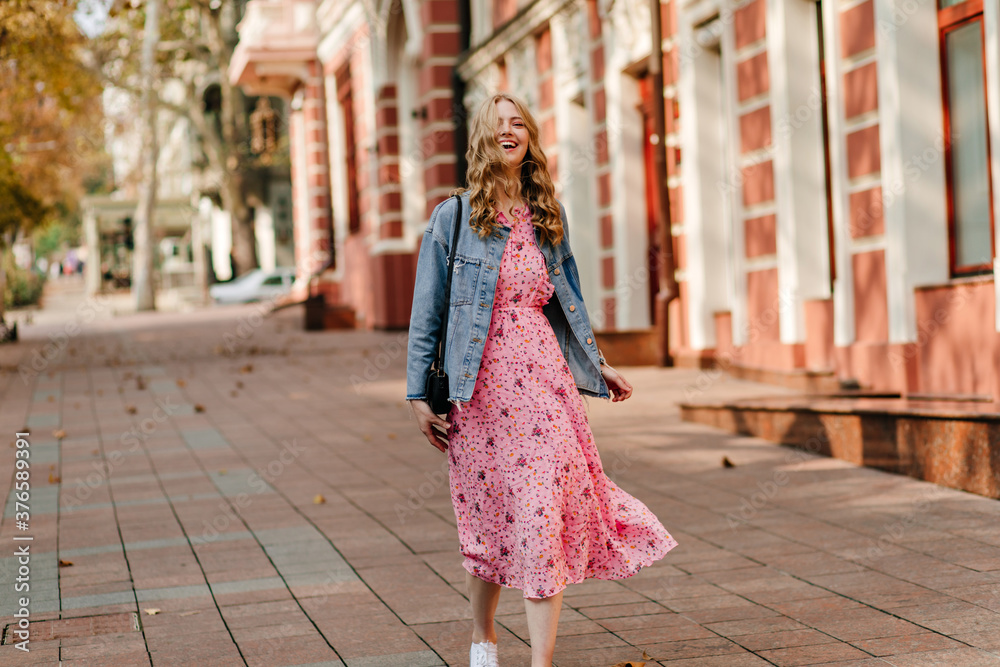 smiling curly blonde goes down the street in a denim jacket and a light pink full-length sundress against the background of the building