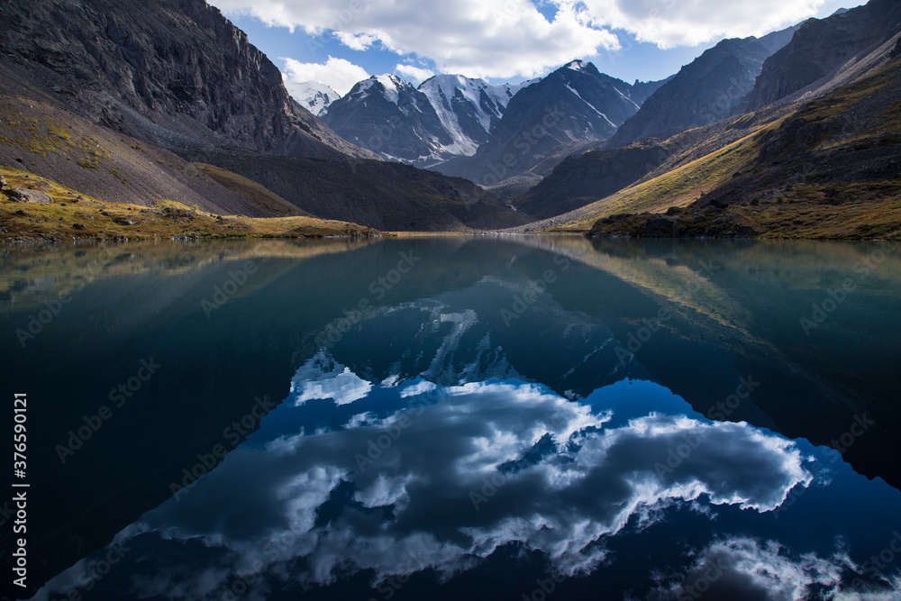 Picturesque clouds are reflected in mounting lake still water. Contrastly lightened mountain slopes on left and right. Snow-capped peaks in the background.