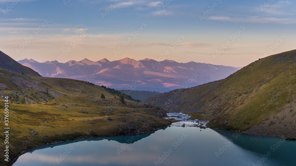 Golden hour sunset in the mountains. There is a lake in the foreground with a river flowing out of it. Distant mountains lightened by the setting sun are in the background. Golden hour sunset in the mountains. There is a lake in the foreground with a river flowing out of it. Distant mountains lightened by the setting sun are in the background.