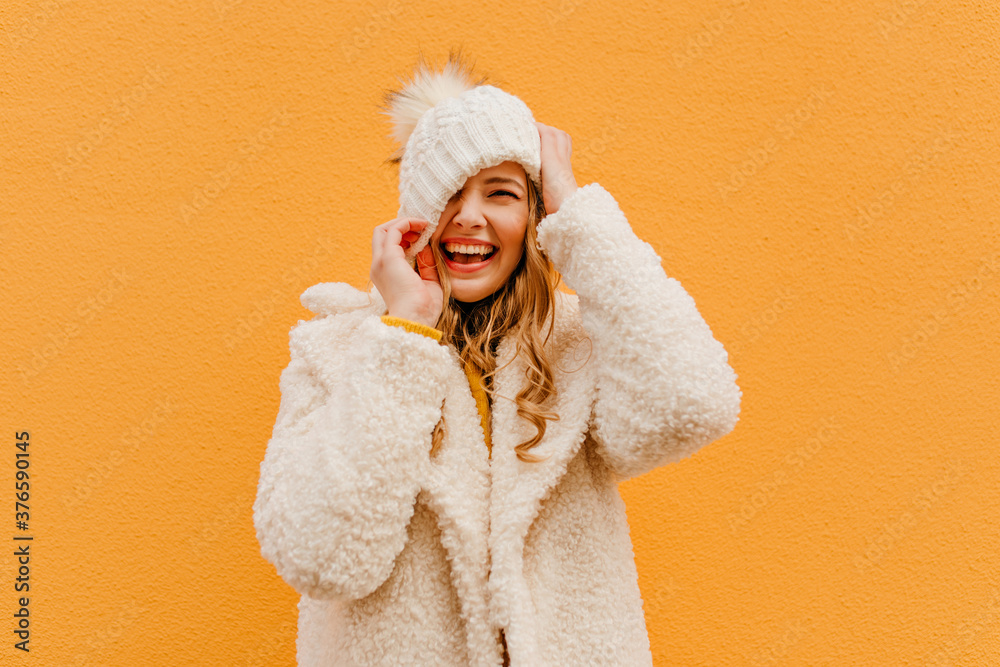 Portrait of girl having fun on isolated background in woolen coat. Blonde puts hat on face - obrazy, fototapety, plakaty 
