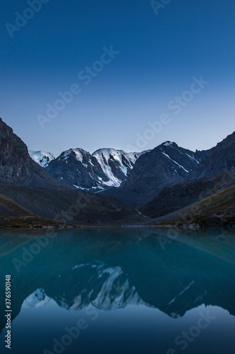 Blue hour in mountains with reflection in a lake. The main subject is a snow-capped mountain in the centre of a frame. The upper half of the frame is a darkening clear blue sky. Portrait orientation.