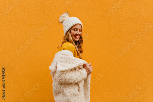 Beautiful curly blonde in orange sweater puts on eco fur coat and looks down flirty photo