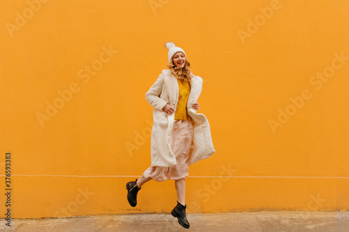 Happy emotional blonde in warm cozy coat and white hat moving down the street across an orange wall