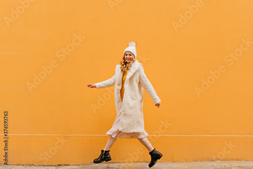 Full-length photo of a beautiful blonde in motion against the background of an orange wall. Photo outside in a warm coat