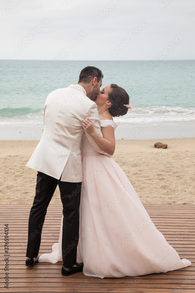 Latino Bride and groom with white tuxedo kissing at the beach