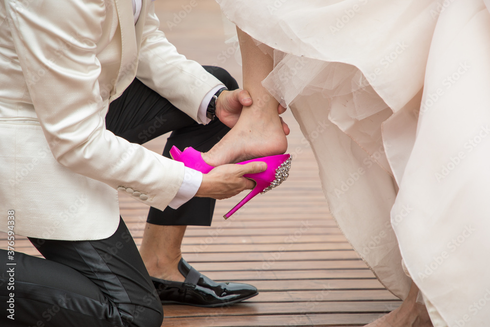 Latino groom with white tuxedo helping bride with her fuchsia color shoe