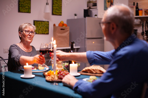 Senior couple clinking glasses with red wine during evening in kitchen. Elderly couple sitting at the table in kitchen, talking, enjoying the meal, celebrating their anniversary in the dining room.