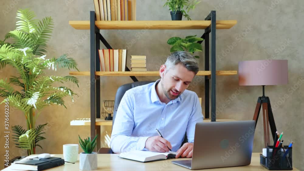 Smiling man at home office study remotely online video call conference with teacher, distance education. Gray-haired bearded guy in wireless earphones looks laptop computer writes information notebook Smiling man at home office study remotely online video call conference with teacher, distance education. Gray-haired bearded guy in wireless earphones looks laptop computer writes information notebook