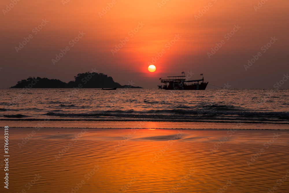 Colorful sunset on a tropical beach. The sun and sky are reflecting in the water. Silhouettes of a boat and a couple of islands are in the background.