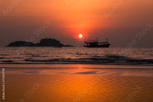 Colorful sunset on a tropical beach. The sun and sky are reflecting in the water. Silhouettes of a boat and a couple of islands are in the background.