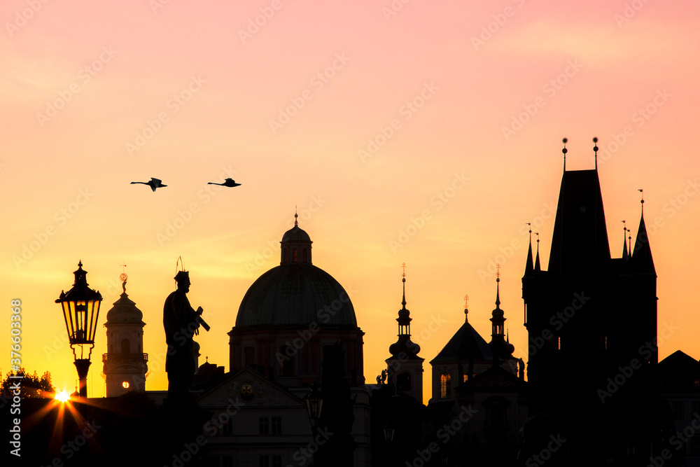 Prague cityscape at sunrise showing silhouettes of buildings, lanterns, statues against the colourful sky and a couple of flying swans 