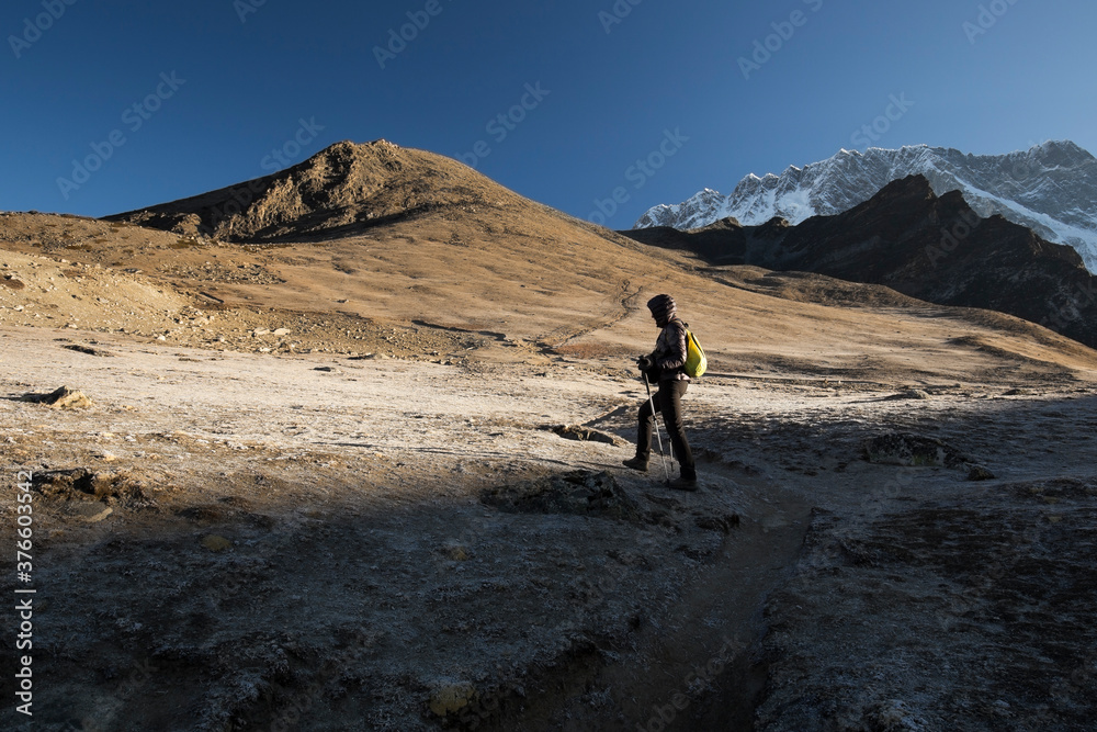 A female trekker with a backpack is going uphill out of the shadow of a mountain into the sunlight. Trekkers' path leads to the top of a hill. Show-capped Mt Lhotse on the right. Bright blue sky