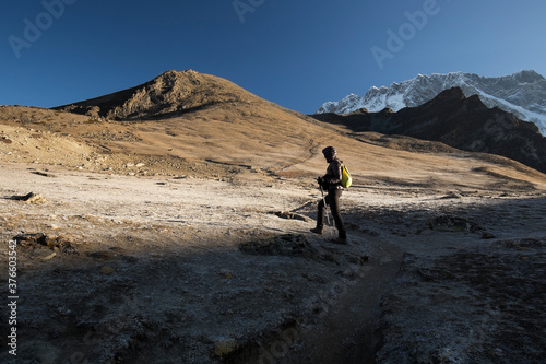 A female trekker with a backpack is going uphill out of the shadow of a mountain into the sunlight. Trekkers' path leads to the top of a hill. Show-capped Mt Lhotse on the right. Bright blue sky