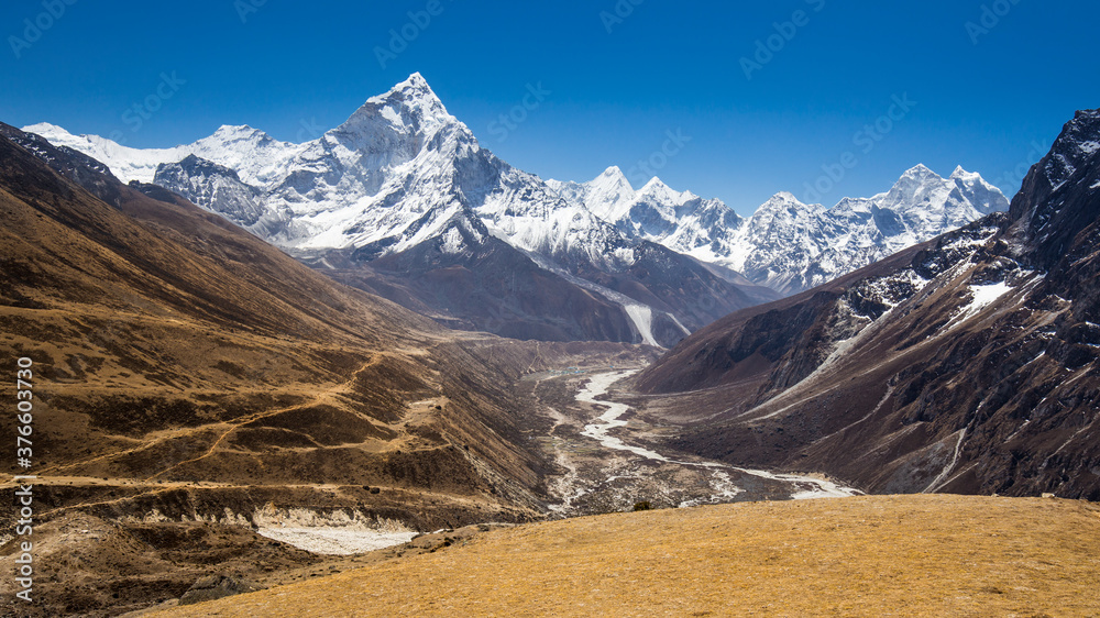 Snow-capped Mt Ama Dablam panorama in Himalayas. View through a valley with a winding river. A black rock on the right. A trekkers' path on a terraced moraine on the left.