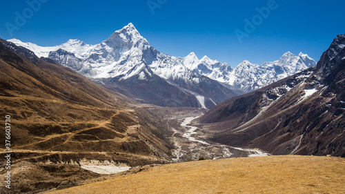 Snow-capped Mt Ama Dablam panorama in Himalayas. View through a valley with a winding river. A black rock on the right. A trekkers' path on a terraced moraine on the left.