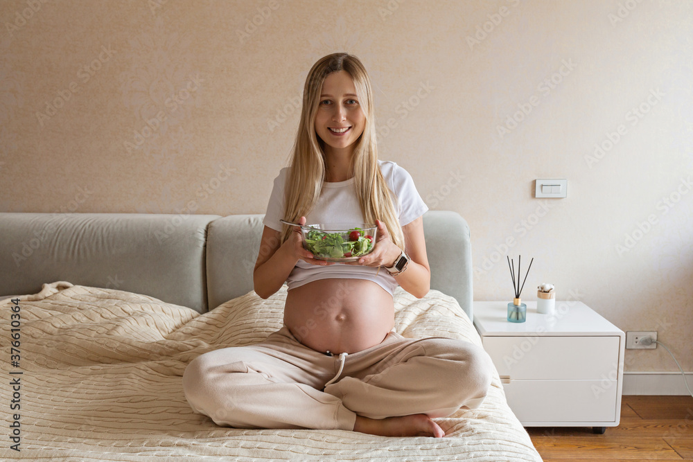 Happy young pregnant woman eating vegetable salad at home. Healthy nutrition and pregnancy concept. Expecting mother holding bowl with fresh cucumbers and tomatoes Happy young pregnant woman eating vegetable salad at home. Healthy nutrition and pregnancy concept. Expecting mother holding bowl with fresh cucumbers and tomatoes