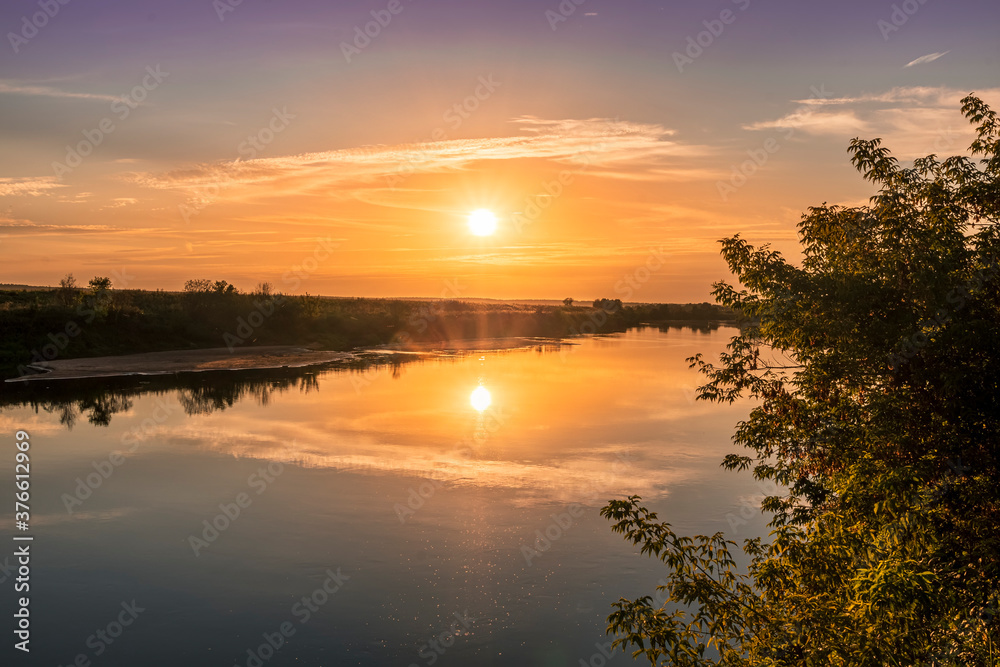 Obraz premium Scenic view at beautiful summer river sunset with reflection on water with green bushes, grass, golden sun rays, calm water ,deep blue cloudy sky and glow on a background, spring evening landscape