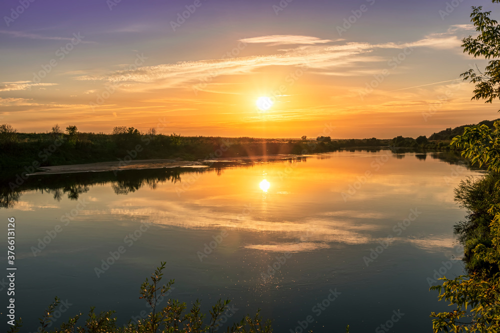 Scenic view at beautiful summer river sunset with reflection on water with green bushes, grass, golden sun rays, calm water ,deep blue cloudy sky and glow on a background, spring evening landscape