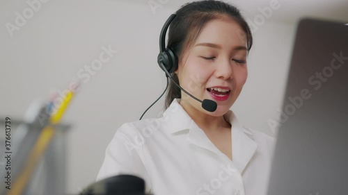 Smiling Asian businesswoman consultant wearing microphone headset of customer support phone operator at workplace.