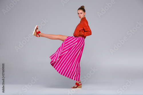 Young woman posing in full growth. Clothes - a long red skirt with a wide white stripe, and orange bomber jacket, wedge sandals. She makes a high leg swing.