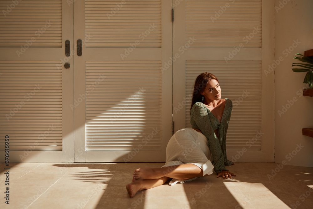Young beautiful girl posing sitting on the floor lit by the bright sun in front of white wall, ook at the camera Young beautiful girl posing sitting on the floor lit by the bright sun in front of white wall, ook at the camera