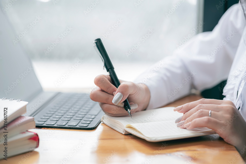 Cropped shot of female hand using pen writing on notebook while sitting at the table in contemporary office room. Cropped shot of female hand using pen writing on notebook while sitting at the table in contemporary office room.