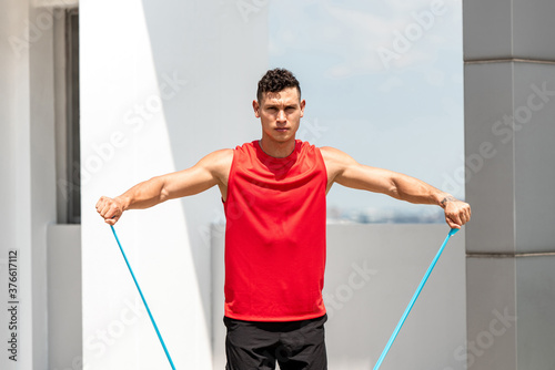 Handsome sports man doing shoulder lateral raise exercise with resistance band outdoors on rooftop in the sun