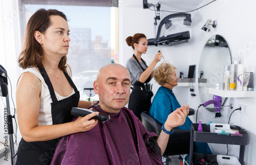 Professional hairdresser woman doing styling of bald man with electric trimmer at barber shop..