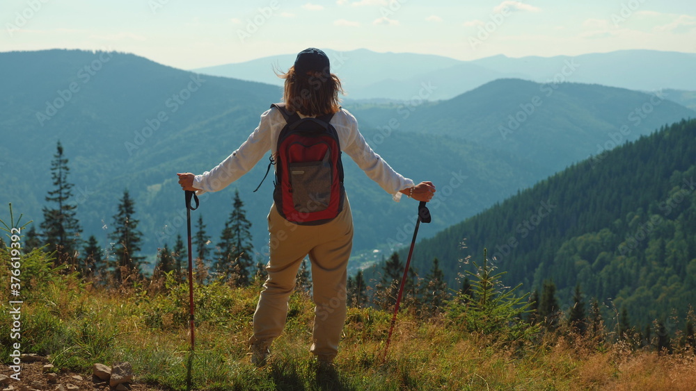 Rear view of an active tourist with trekking poles and a backpack enjoying the landscape while standing on the top of the mountain