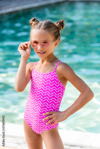 child in swimsuit touching sunglasses while posing with hand on hip near pool