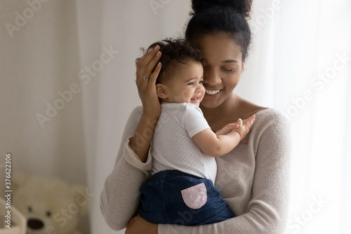 Front view affectionate african american mum holding little funny mixed race toddler daughter or son, enjoying spending daycare time together in baby room, showing love and devotion to small kid.