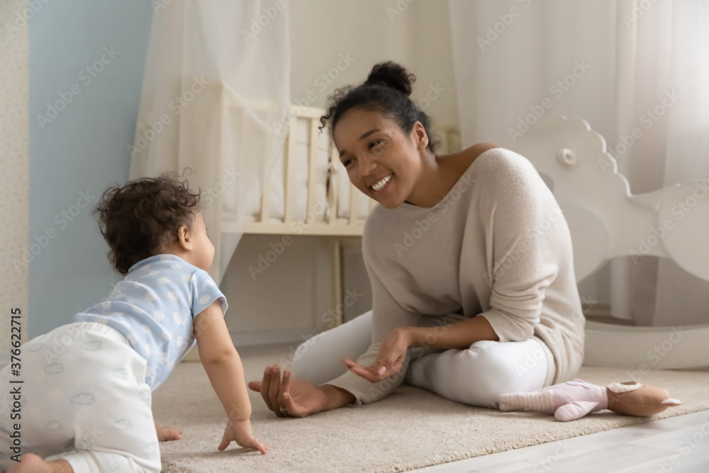 Adorable happy mixed race baby girl or boy creeping, walking on knees and hands on carpet floor to smiling young african american mother, having fun together in children bedroom, childcare concept. Adorable happy mixed race baby girl or boy creeping, walking on knees and hands on carpet floor to smiling young african american mother, having fun together in children bedroom, childcare concept.