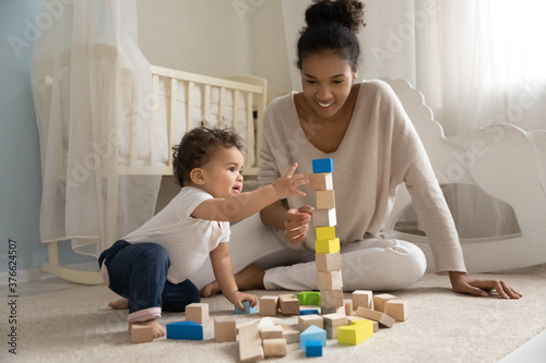 Happy young african american mother or nanny sitting on floor carpet, constructing building with wooden cubes with curious adorable small toddler baby boy girl in children room, childcare concept. photo