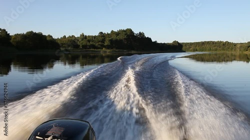 Motor boat with an powerful outboard motor fast gliding on river calm water, beautiful view from stern on transom Wake at Sunny summer day on trees on shore and blue sky background, boating lifestyle