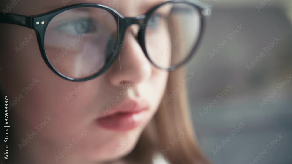 A close-up shot of a serious little 8 years old girl in glasses looking to a laptop screen. Remote education technologies and homework. A close-up shot of a serious little 8 years old girl in glasses looking to a laptop screen. Remote education technologies and homework.