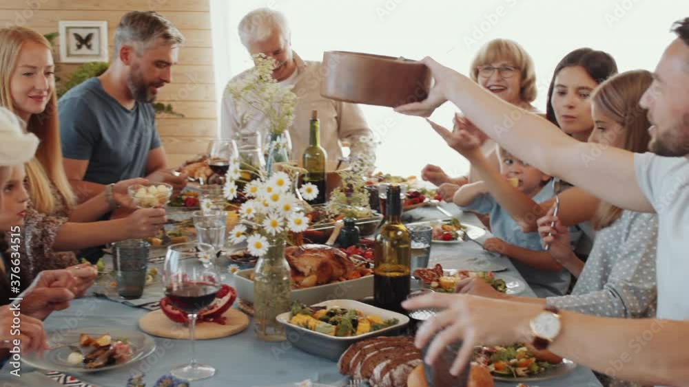 Handheld camera shot of happy large family having holiday dinner together: senior grandfather talking to children while others eating delicious food and passing dish