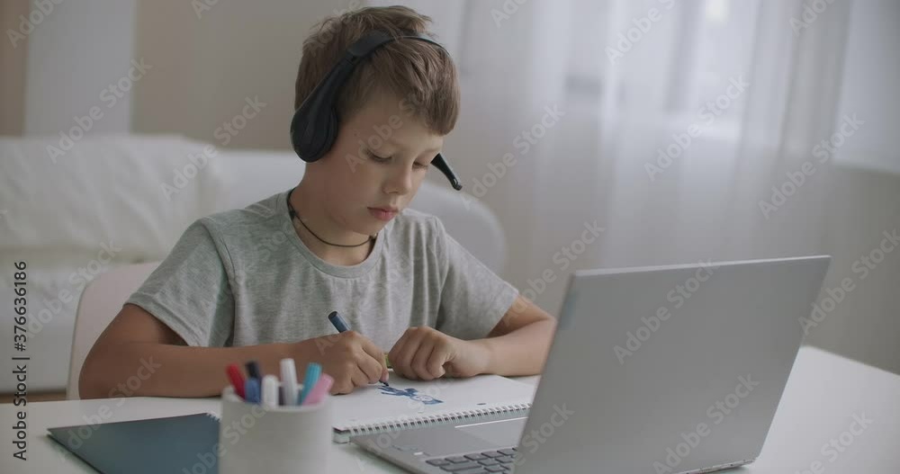 homeschooling at pandemic of coronavirus, little boy is listening teacher by headphones and using laptop, drawing on paper homeschooling at pandemic of coronavirus, little boy is listening teacher by headphones and using laptop, drawing on paper