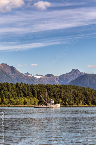 Shot of a commercial fishing boat anchored in a harbour in Sitka, AK. Green forest, mountains, and gorgeous blue sky in the background. Vertical shot.