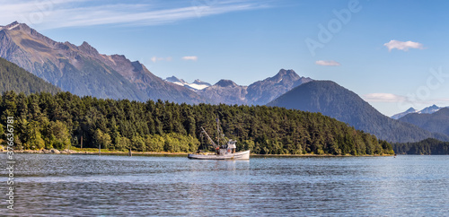 Beautiful panoramic shot of a commercial fishing boat anchored in a harbour in Sitka, AK. Green forest, mountains with snowy peaks, and gorgeous blue sky in the background.