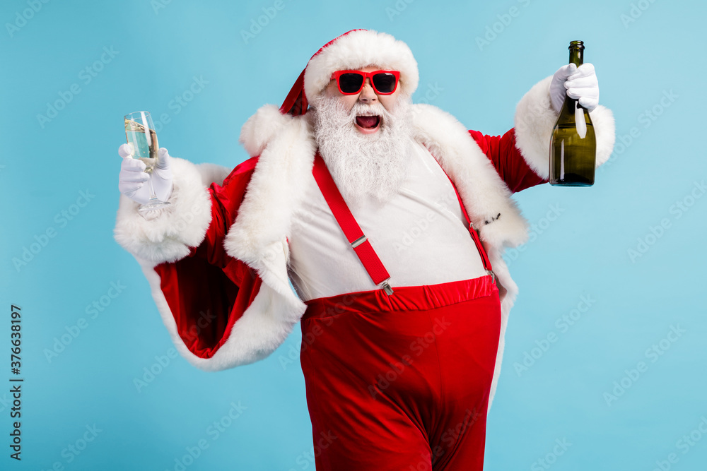 © deagreez - Photo of retired old man grey beard funky hold bottle full glass golden wine drink having fun open mouth wear santa x-mas costume suspender sunglass cap isolated blue color background