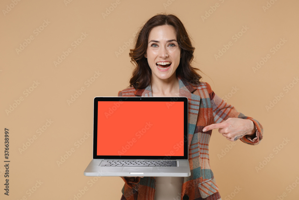 Funny young brunette woman 20s in casual checkered jacket pointing index finger on laptop pc computer with blank empty screen mock up copy space isolated on beige colour background, studio portrait. Funny young brunette woman 20s in casual checkered jacket pointing index finger on laptop pc computer with blank empty screen mock up copy space isolated on beige colour background, studio portrait.