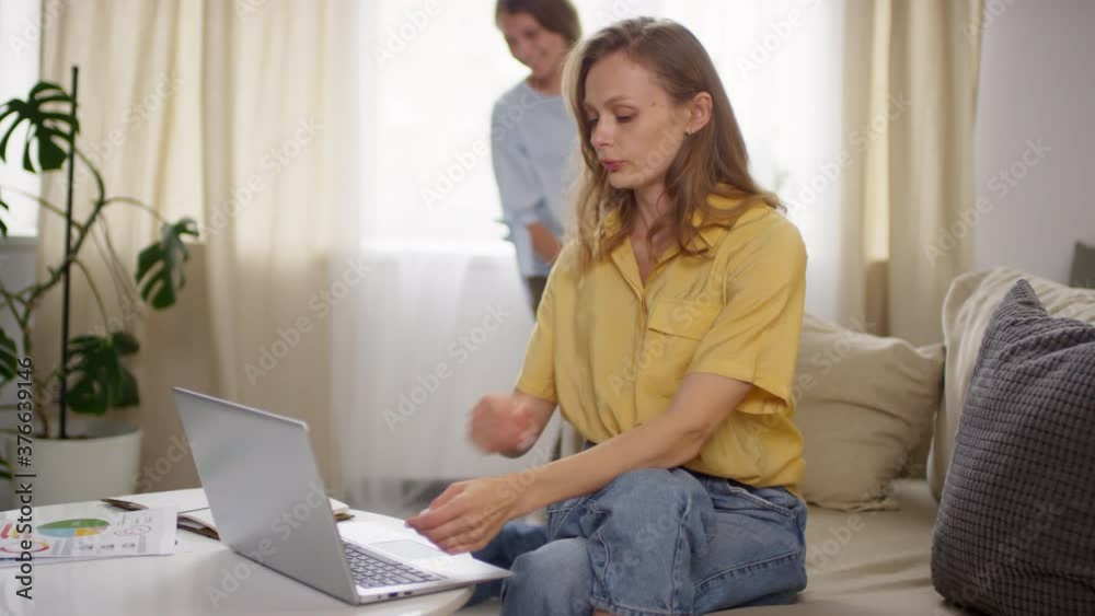 Handheld shot of beautiful young woman sitting on couch and grabbing her head in frustration while talking and working on laptop. Happy teenage girl playing with little brother in background Handheld shot of beautiful young woman sitting on couch and grabbing her head in frustration while talking and working on laptop. Happy teenage girl playing with little brother in background
