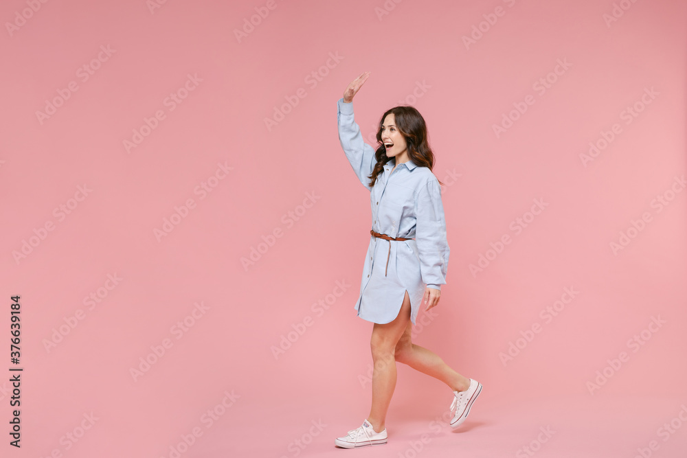 Full length side view portrait of funny young brunette woman 20s wearing casual blue shirt dress waving and greeting with hand as notices someone isolated on pastel pink colour background in studio. Full length side view portrait of funny young brunette woman 20s wearing casual blue shirt dress waving and greeting with hand as notices someone isolated on pastel pink colour background in studio.