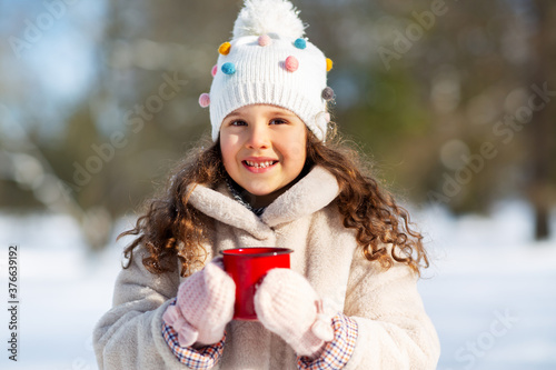 childhood, christmas and season concept - happy little girl with cup of hot tea in winter park
